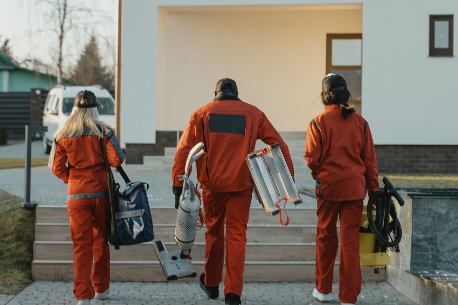 Tre servicemedarbejdere i orange arbejdstøj går op mod et hus med støvsuger og rengøringsudstyr
